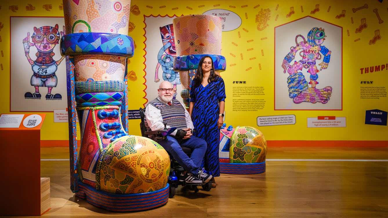 A photo of a man with a white beard and thick framed glasses seated in a wheel chair next to a woman wearing a blue patterned jumpsuit. They are next to a brightly coloured large sculpture of legs