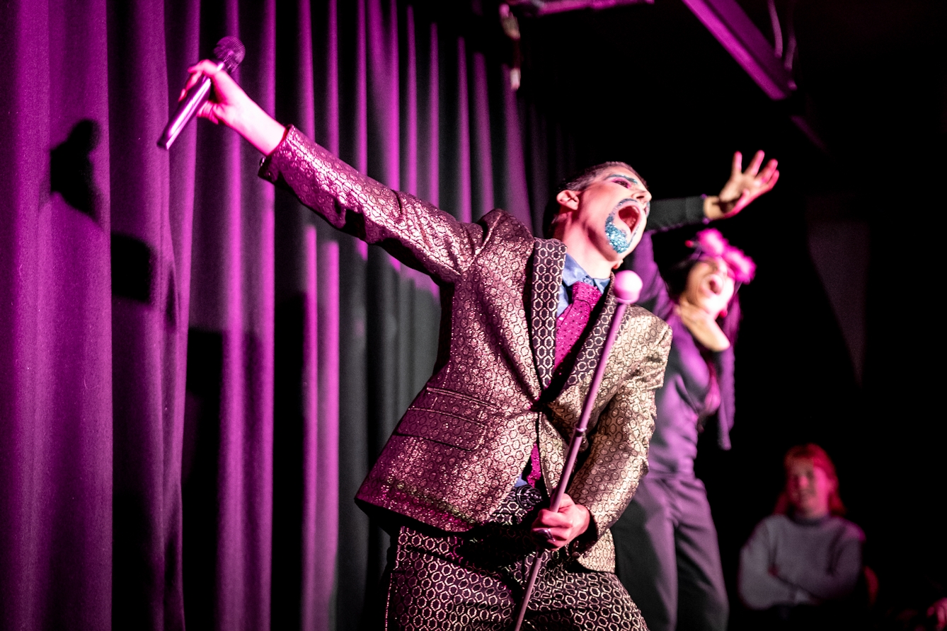 Photograph showing a performer on a stage in a small theatre. The performer is dressed in a shiny gold suit. Their right hand is outstretched, holding a microphone. Their mouth is open in speech or song. In their left hand they are holding a white cane.