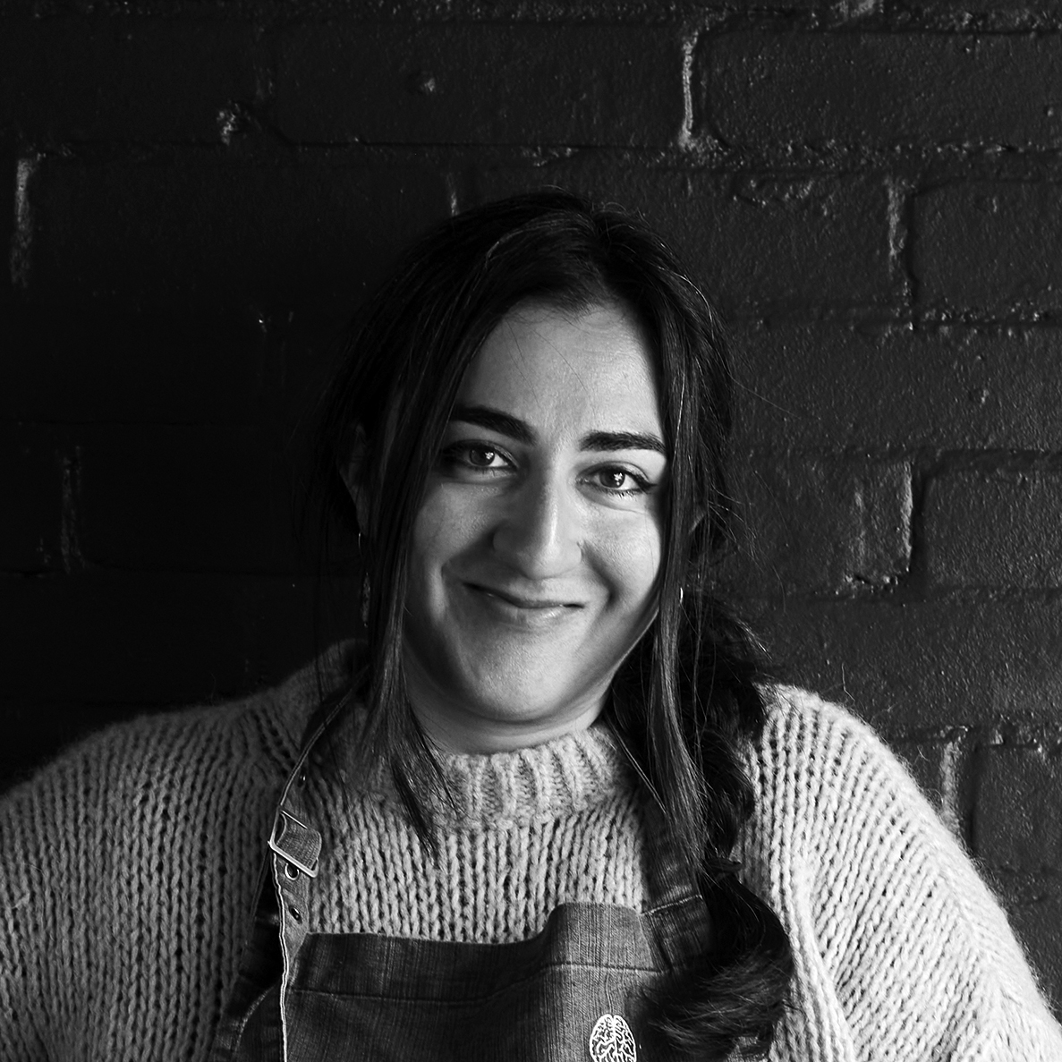 Head and shoulders photograph of a woman with long dark hair wearing a woollen jumper and an apron
