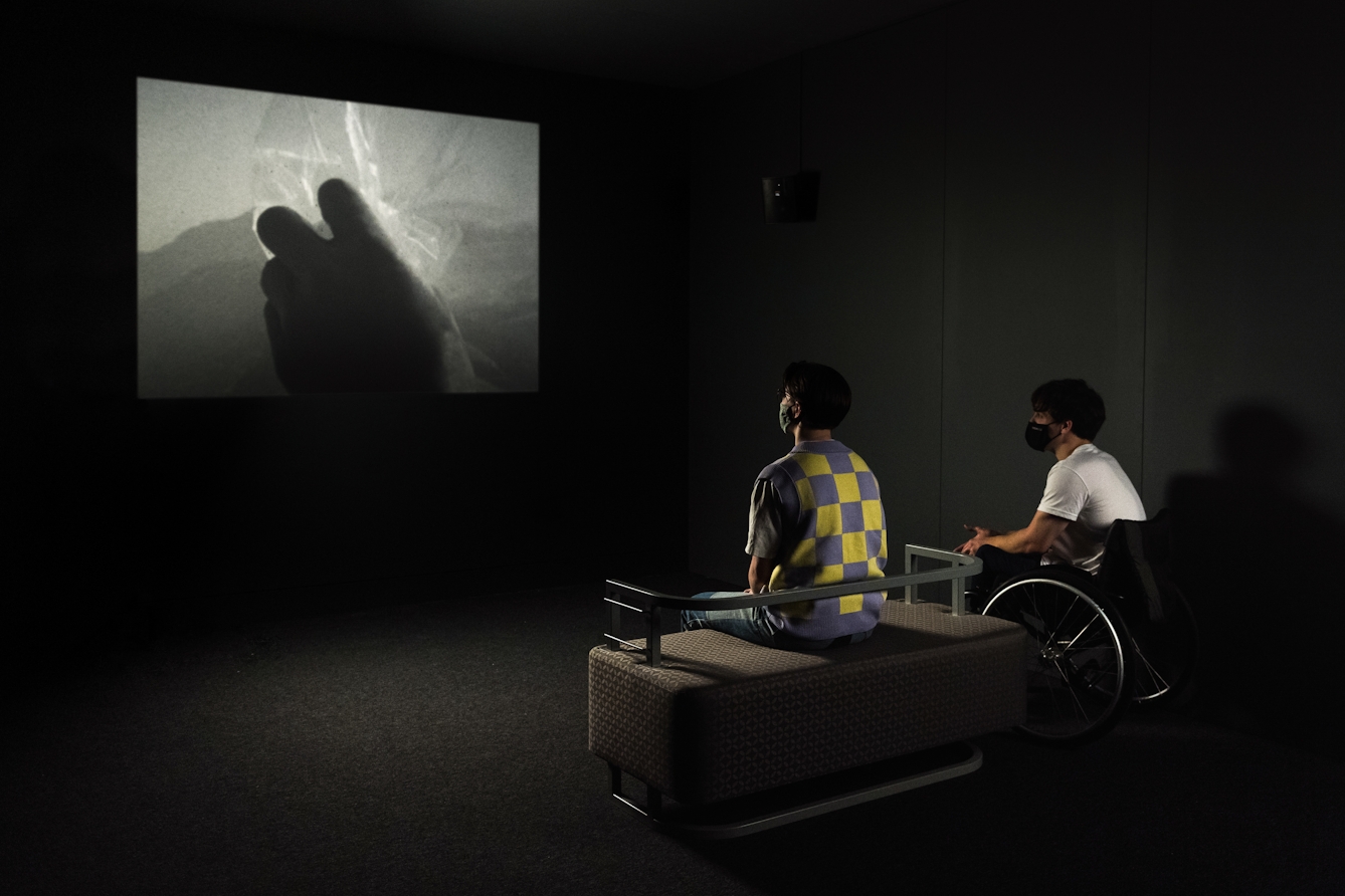 Colour photograph of two young men watching a film on a large screen in a dark room. One of the boys, wearing a yellow and blue checked sweater vest, is seated, whilst the other boy is in a wheelchair. Both are wearing face masks. The film shows the dark silhouette of a hand holding some plastic.  