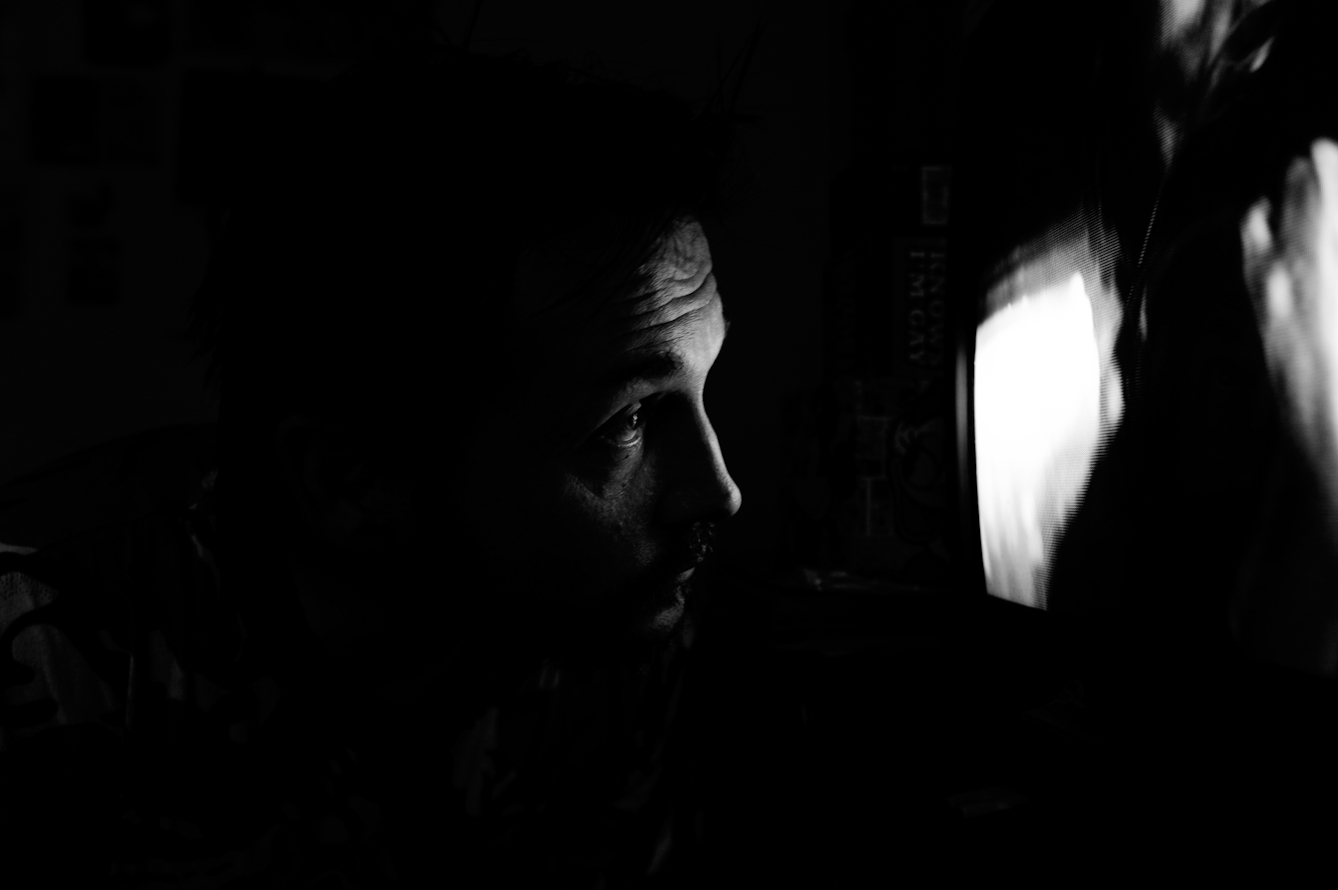 Black and white photograph showing a dark room with a tv screen. The light from the tv screen illuminated the profile of a man's face looking into the screen, several inches from it's surface.