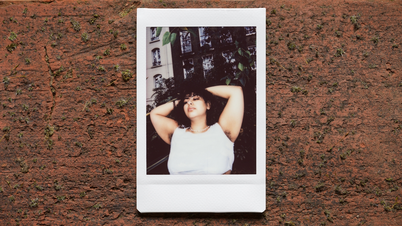 Photograph of an Instax Mini instant film print resting on a textured brick surface.  The print shows a woman against the branches and leaves of a tree outside a tall row of houses, with her arms raised up to show her armpit hair. She is wearing a white top.