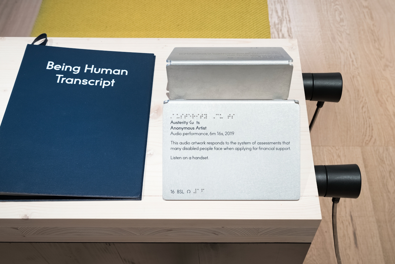 A photograph of the Being Human Transcript, blue with white text, and audio handsets relating to an audio performance titled 'Austerity' by an anonymous artist. The plaque reads 'This audio artwork responds to the system of assessments that many disabled people face when applying for financial support'. The plaque also displays the title in braille and a number relating to BSL captions.
