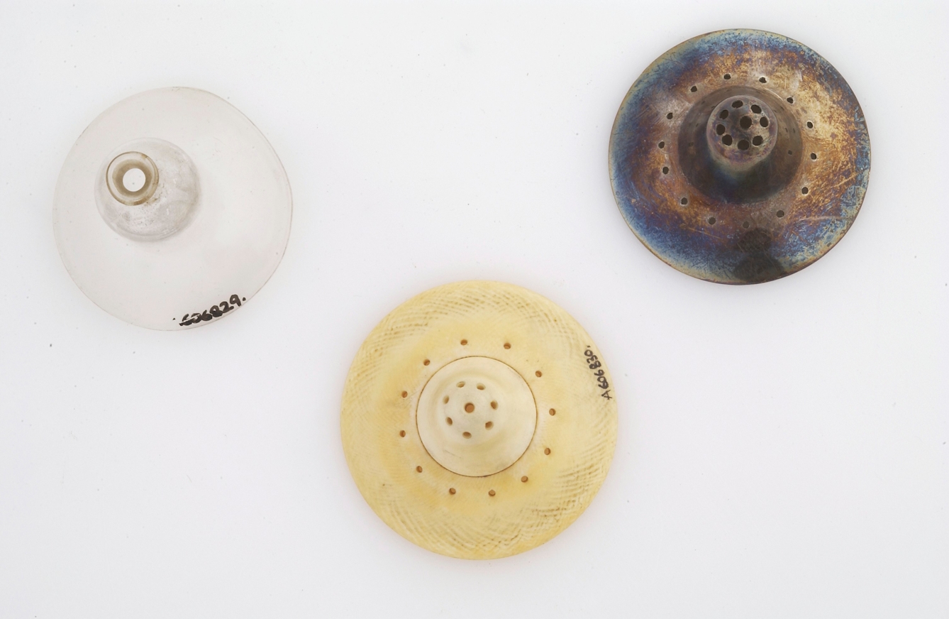 Colour photograph of three nipple shields (objects in the shape of nipples with holes in for feeding through). The one on the left is made of glass, the middle one is ivory, and the one on the right is made of silver.