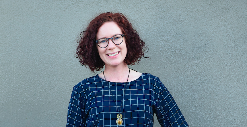 Photo of  a woman. She has curly hair and is wearing glasses. and checked top.