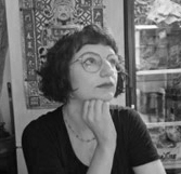 Black and white photograph of the head and shoulders of a young woman with short dark hair and round glasses. The woman is looking up and to the right.