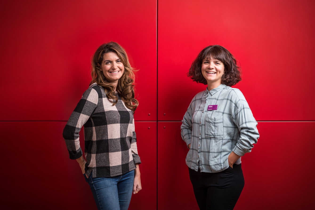 A portrait of two women standing against a bright red background.