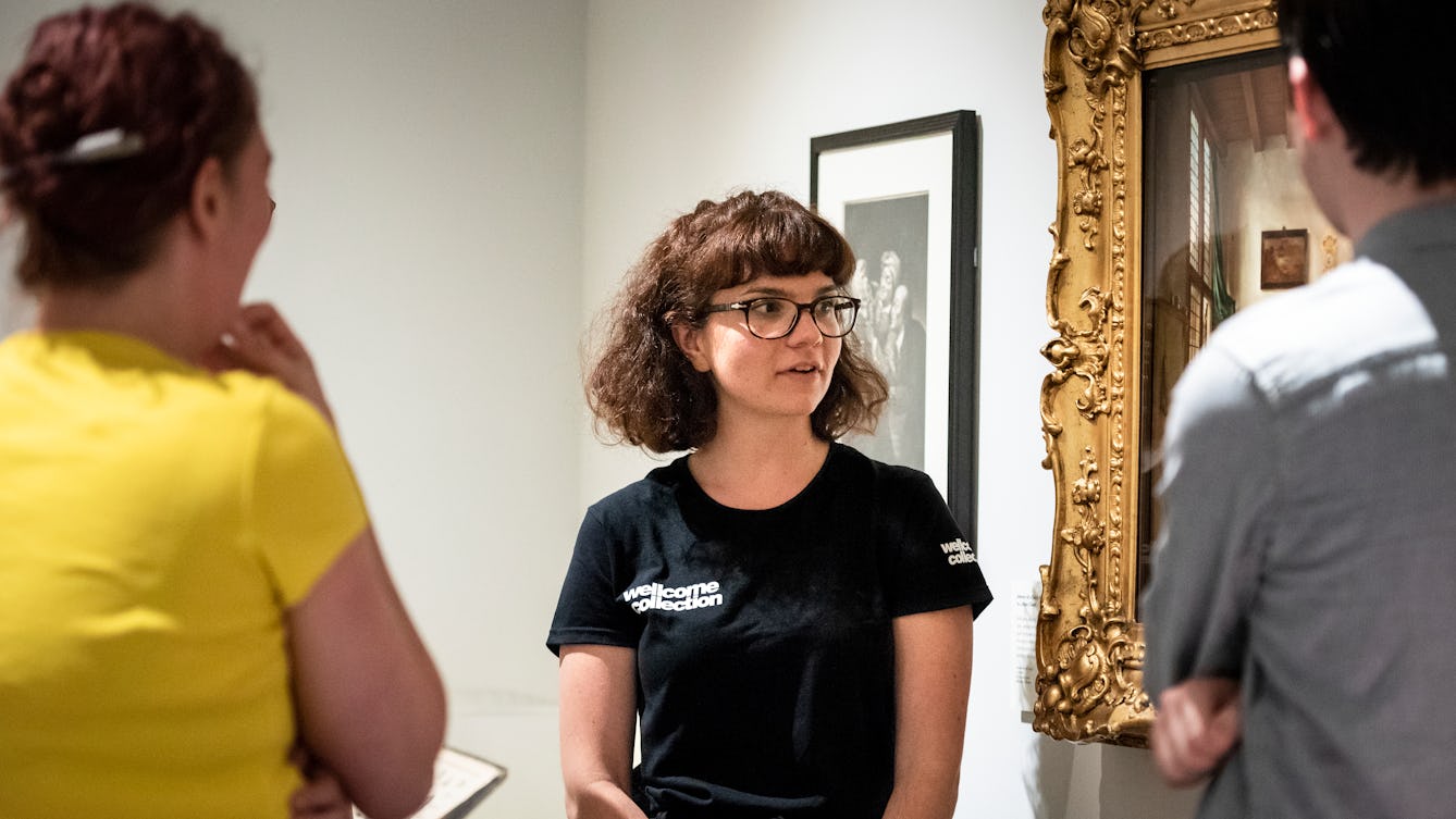 Photograph of a museum gallery visitor experience assistant wearing a black T-shirt with 'Wellcome Collection' written on it, giving a tour of the exhibition space to members of the public. They are looking at a painting, partially in view, which has an ornate gold frame.  