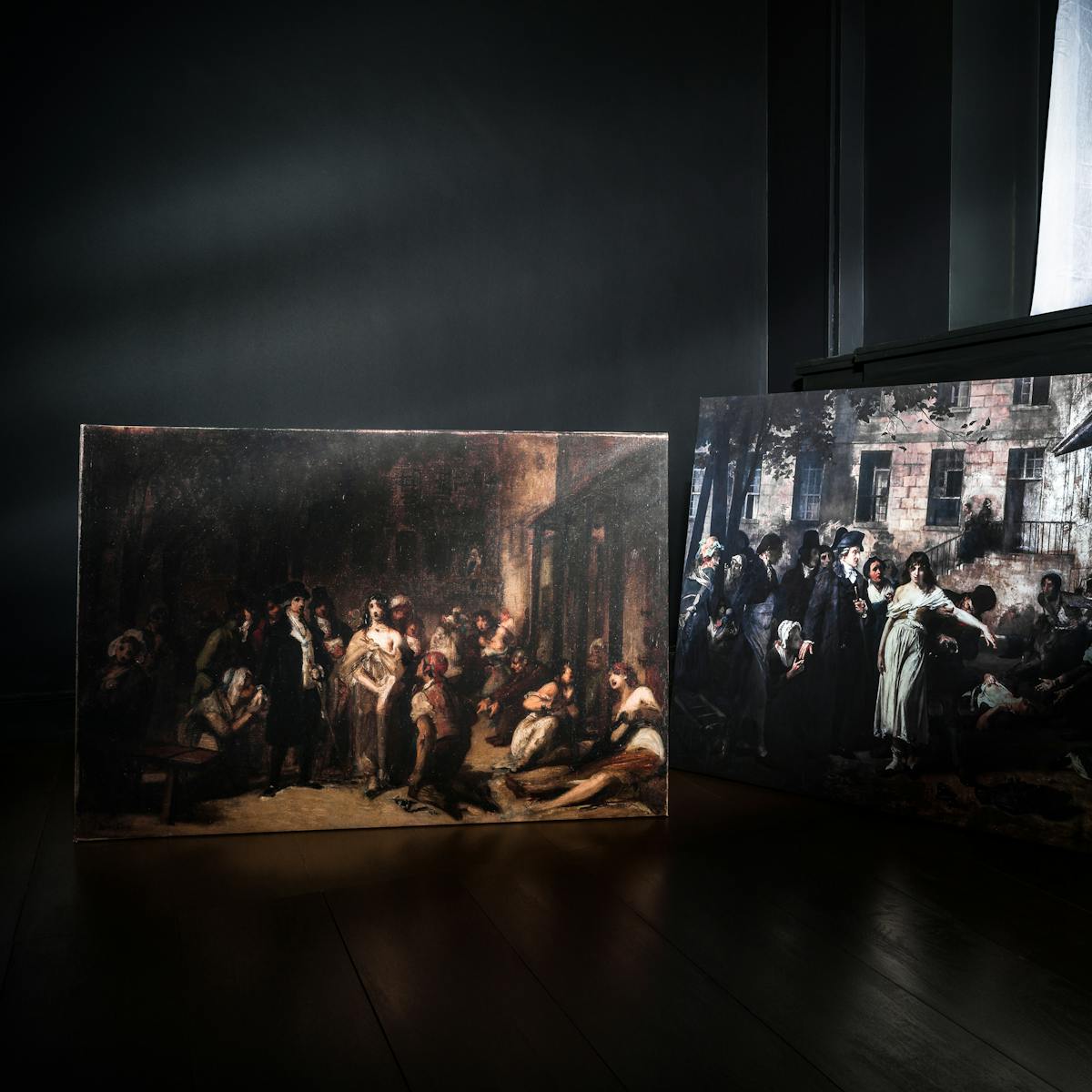 Photograph of two oil paintings propped against the walls in the corner of a dark room, on a wooden floor.  To the right is a recessed window with a white fabric curtain hanging in front of the glass. 