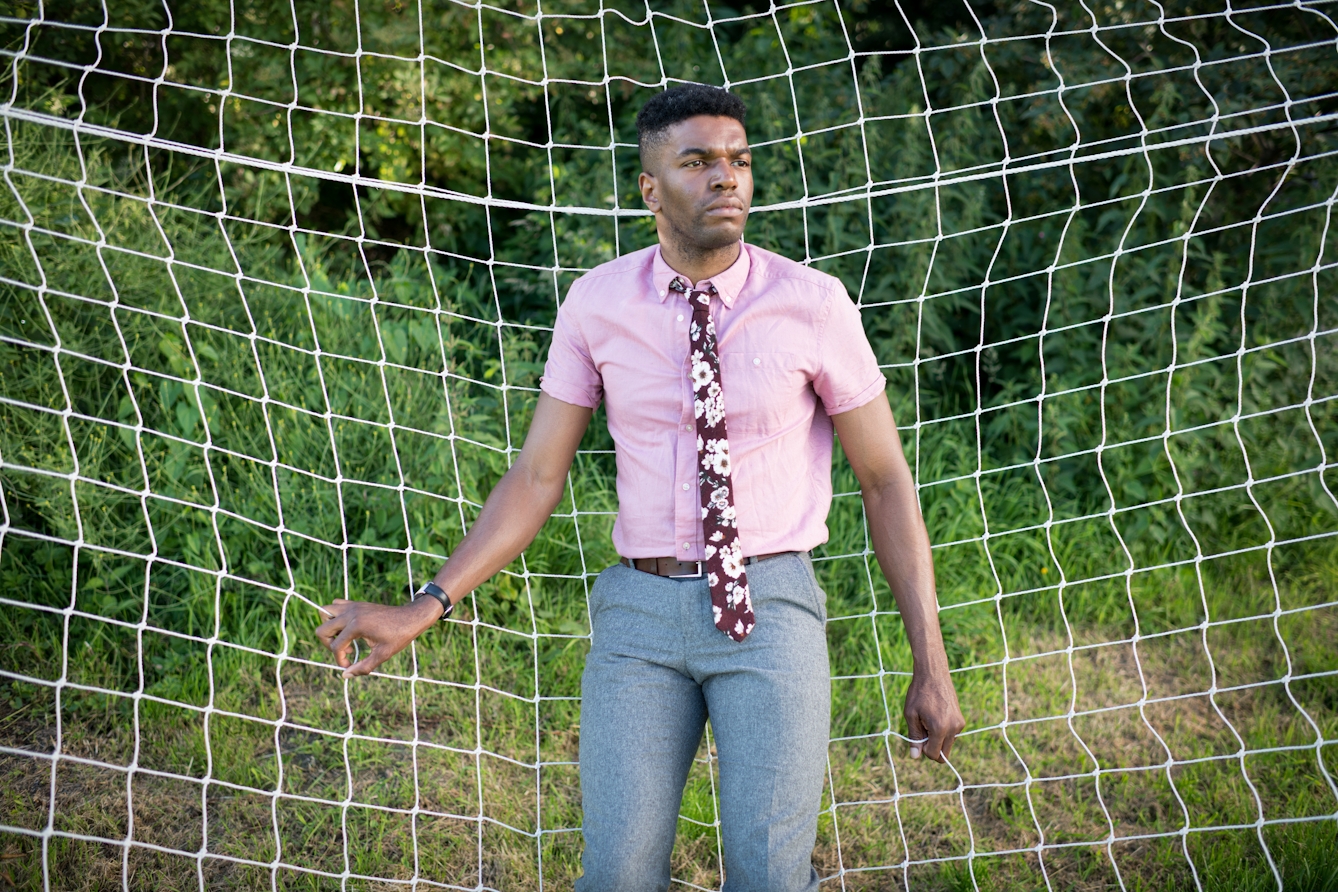 Photograph of a man leaning back against the netting of a football goal. He is gripping the netting with his hands a looking off into the distance. He is wearing a pink short sleeved shirt, floral tie and grey trousers. Behind the goal netting is green foliage.