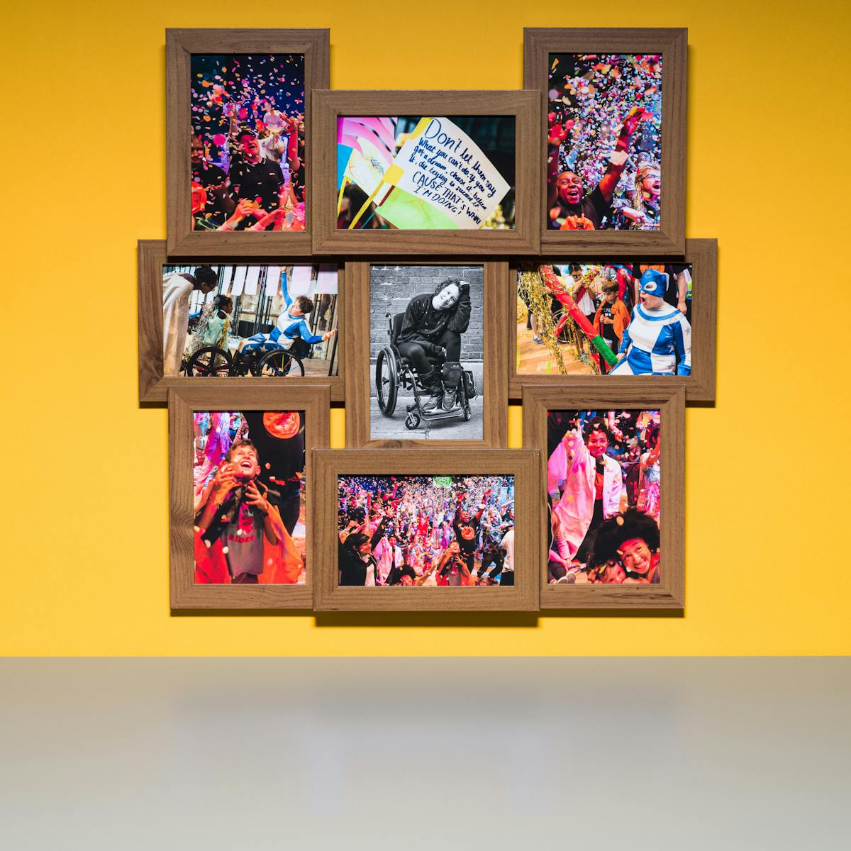 Photograph of a multi-frame photo frame containing nine photographs, one in black and white and eight in colour. The frame is hung on a yellow wall above a grey tabletop.