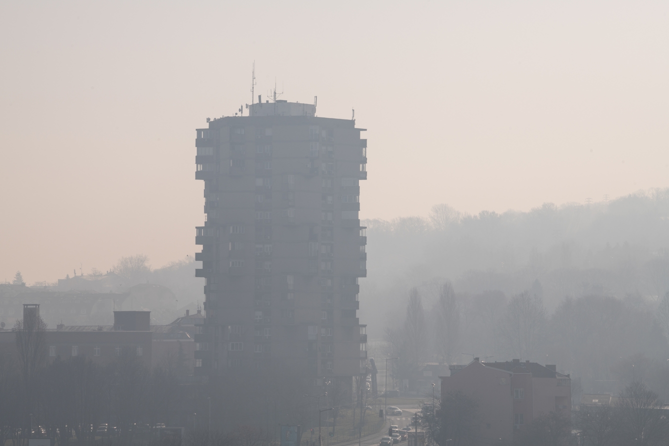 Photograph of a tall high rise residential building complex in the centre. Surrounding the building are other smaller buildings, tree-like shapes and a street with vehicles to the left of it. It is very misty and foggy, and the mist is occluding the buildings. 