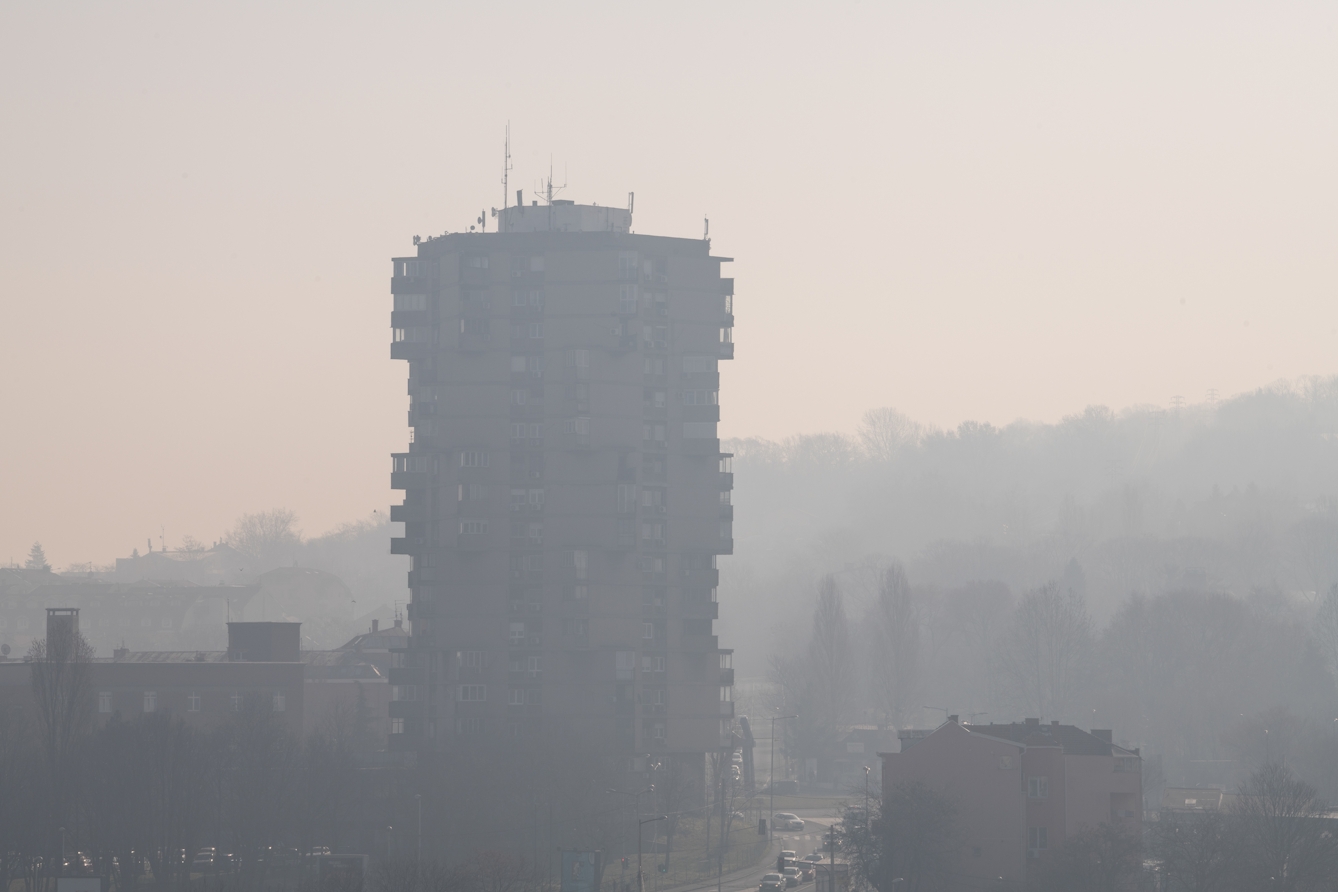 Photograph of a tall high rise residential building complex in the centre. Surrounding the building are other smaller buildings, tree-like shapes and a street with vehicles to the left of it. It is very misty and foggy, and the mist is occluding the buildings.