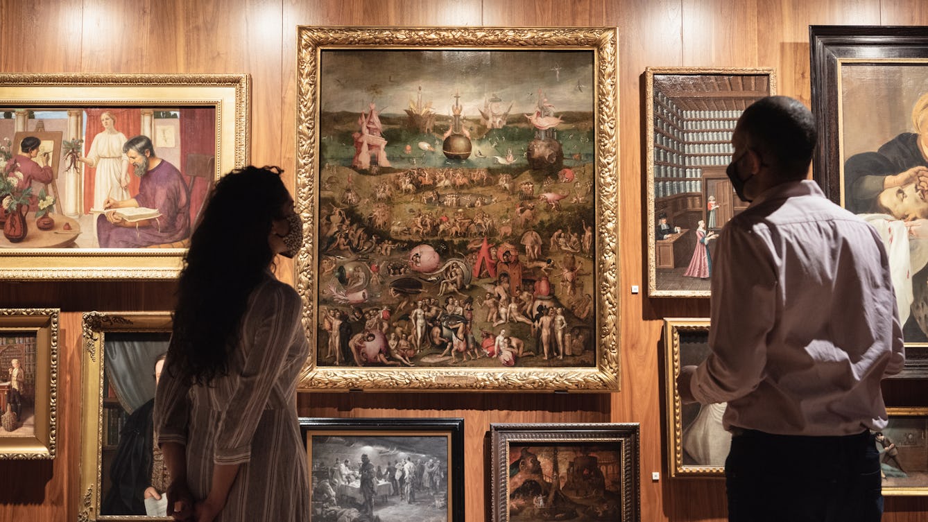 Photograph of two people wearing face coverings looking at an oil painting on the gallery wall in an ornate gold frame. Painting title 'Garden of earthly delights'. The painting is filled with nude figures in a vast garden landscape.