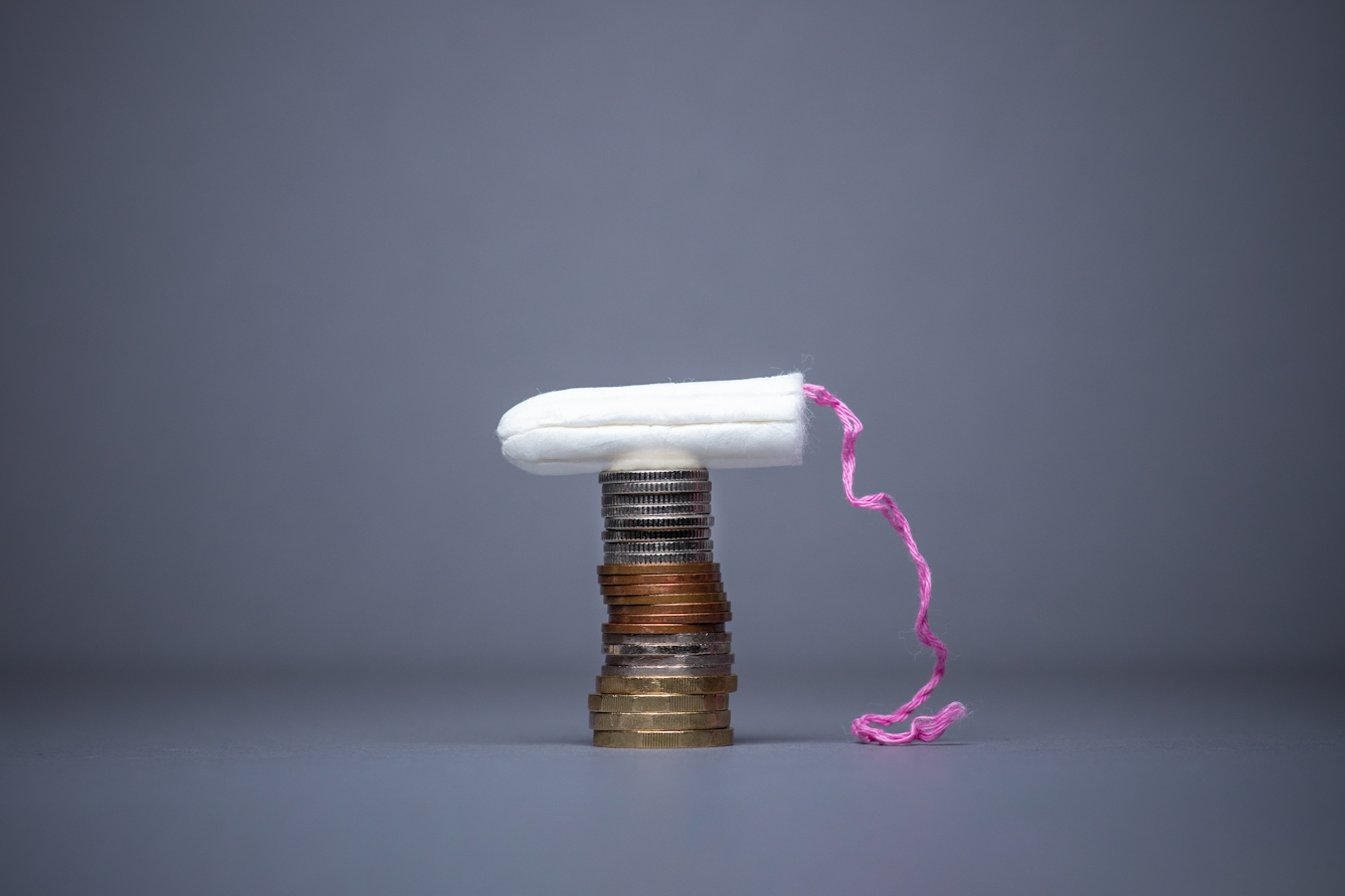 Photograph of an unwrapped tampon resting on top of a stack of British coins. The pink cord can be seem hanging down onto the blue background.