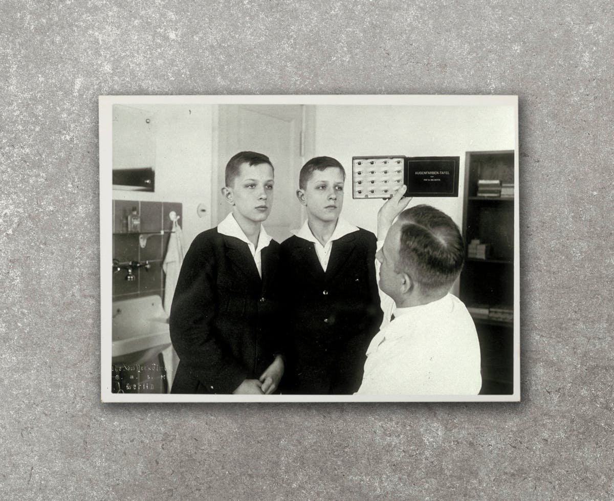 Black and white photograph of two adolescent, identical twin boys standing side by side in front of a man iwth a white coat who holds up a chart with illustrations of different eyes while looking at the eyes of the boys. The three figures are in a laboratory.