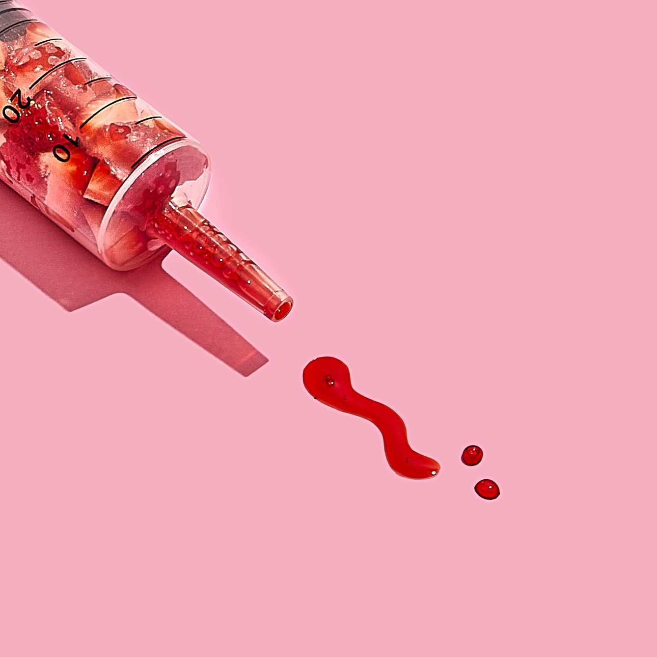 Photograph of a medical syringe lying on its side on a bright pink background. The syringe is full of chunks of cut up strawberry, compressed down into the lower half of the granulated tube. The nozzle of the syringe contains bright red strawberry juice. Some of this juice has been pushed out of the syringe and formed two dots and a squiggle on the background, reminiscent of marks made with a pen on paper.