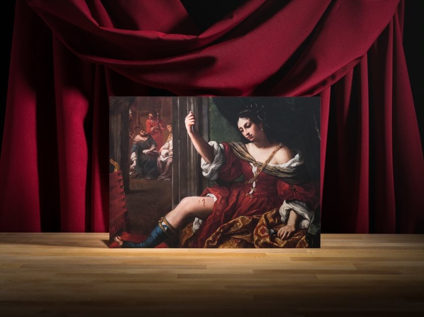 Photograph of an oil on canvas painting of a woman raising her hand and holding up a knife towards her exposed thigh. Her thigh is wounded and blood is coming out of the wounded sight. She is wearing a grand red dress and a grand looking necklace. 