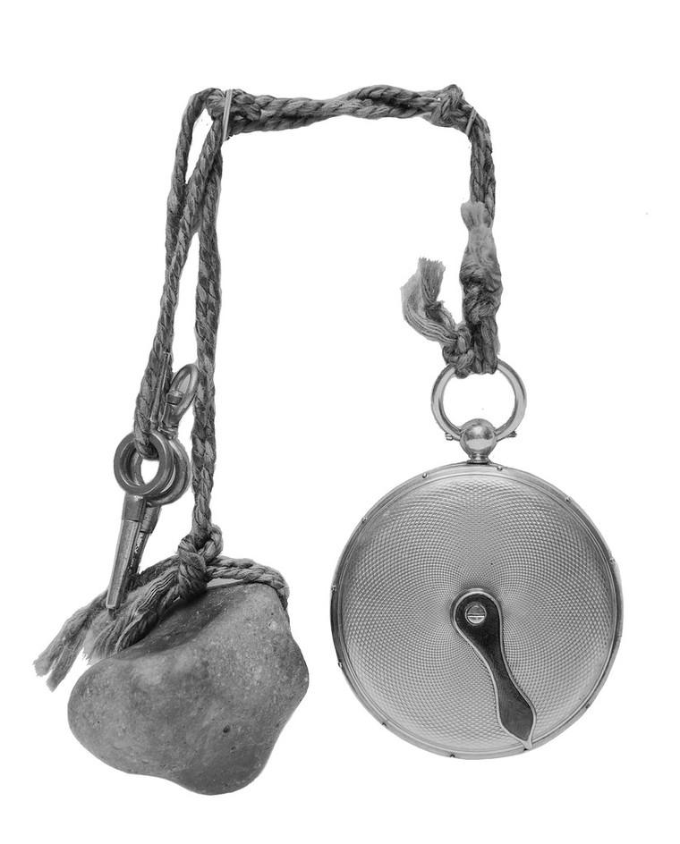 A black and white photograph of a pocket-watch style object. The object consists of a pebble, two small keys and a small round clock, all attached to a length of string. The clock is made of metal, it has a single hand and 12 equally spaced, raised dots around the edge.