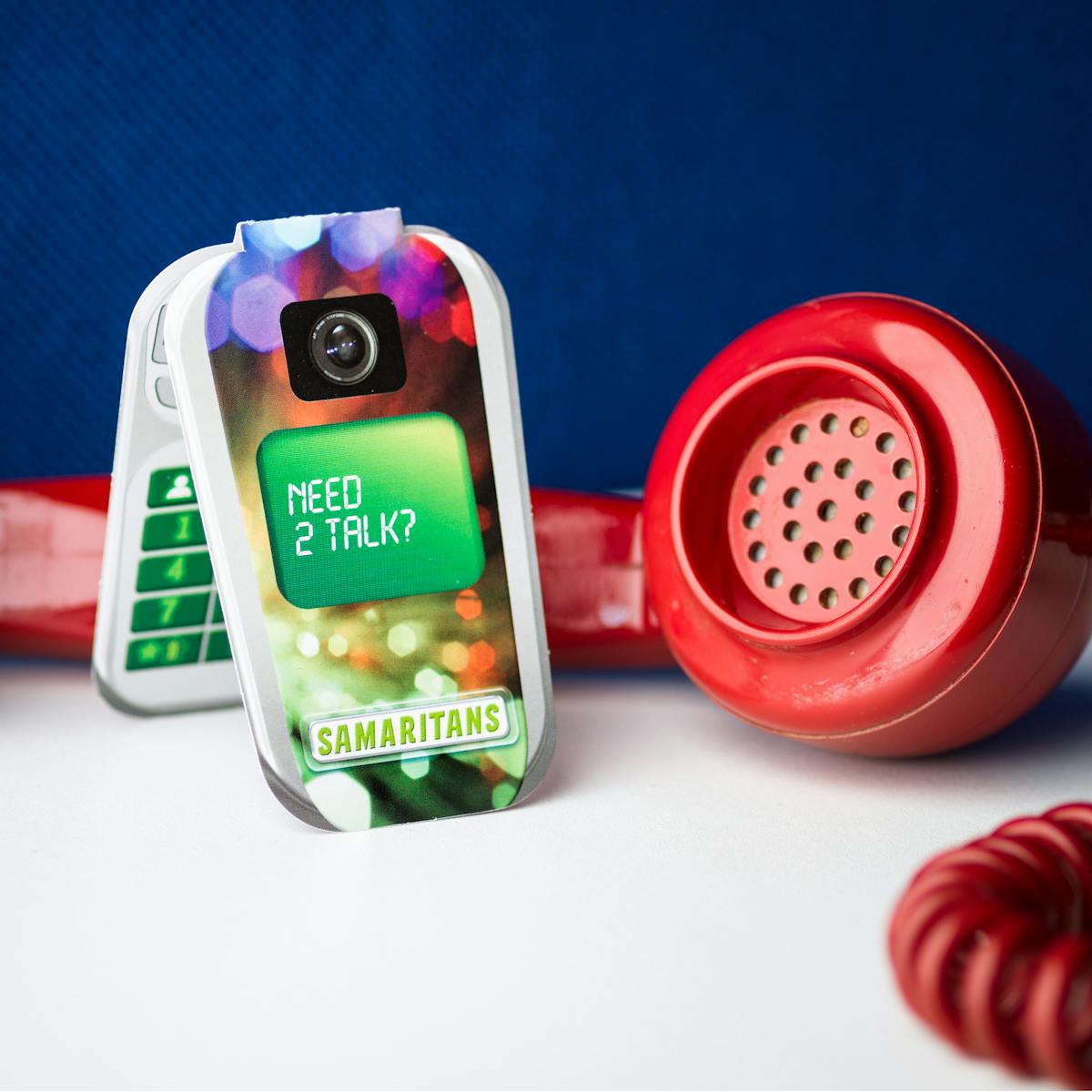 Photograph of an old red rotary phone receiver lying on its side on a white desk. Behind it is a blue felt desk back. between the earpiece and the mouthpiece is a Samaritans counseling service information leaflet in the shape of a flip mobile phone. on the front is written, 'Need 2 talk?'.
