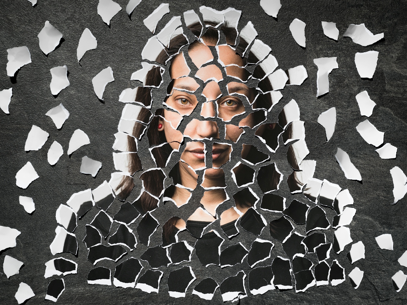 Photograph of a collages of torn up pieces of a photographic print scattered on a grey slate background. There are fragments of eyes, lips, hair and dark and light tones. The arrangement of the fragments are starting to form a recognisable face.