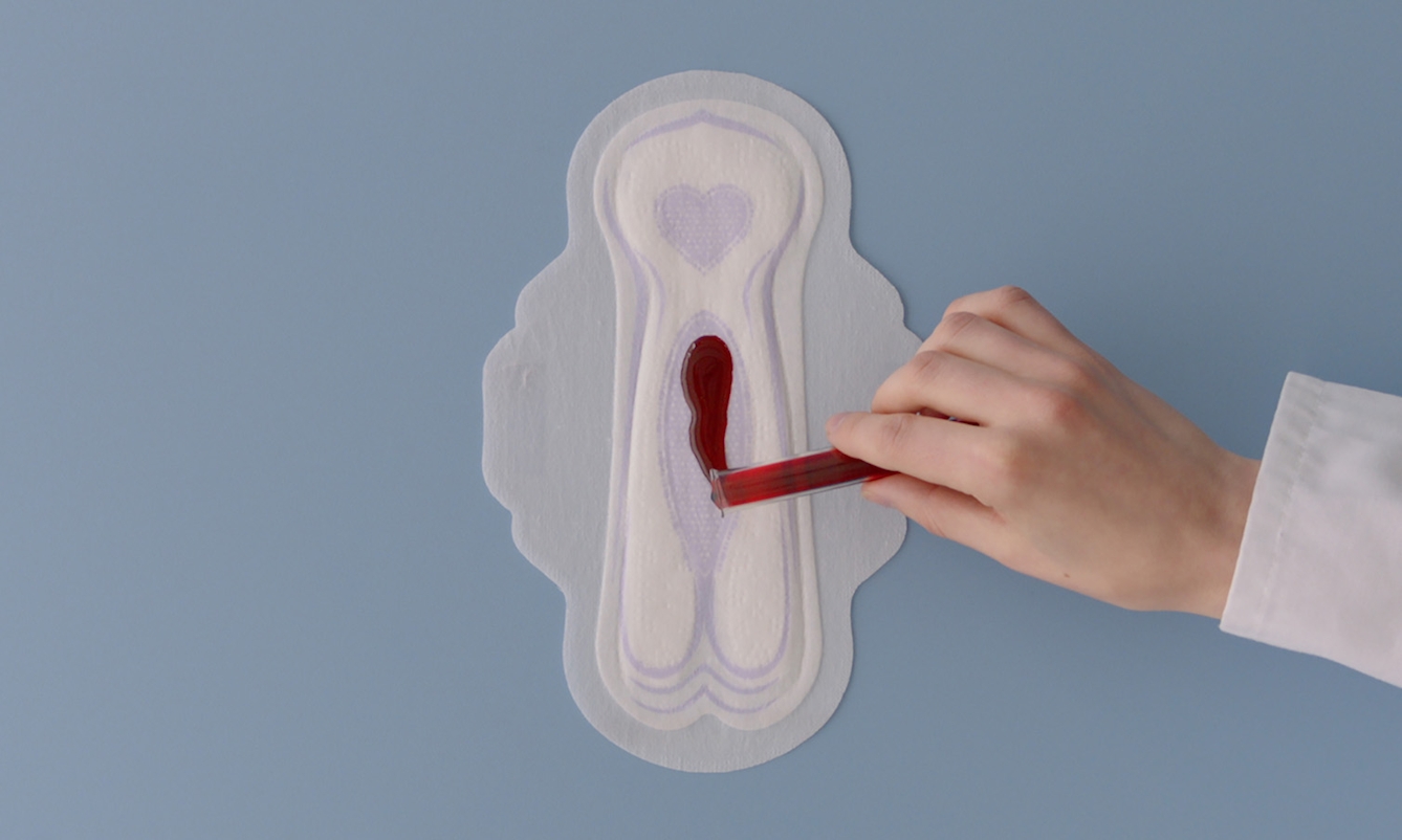 Person in lab coat pours a test tube of red liquid onto a sanitary pad. 