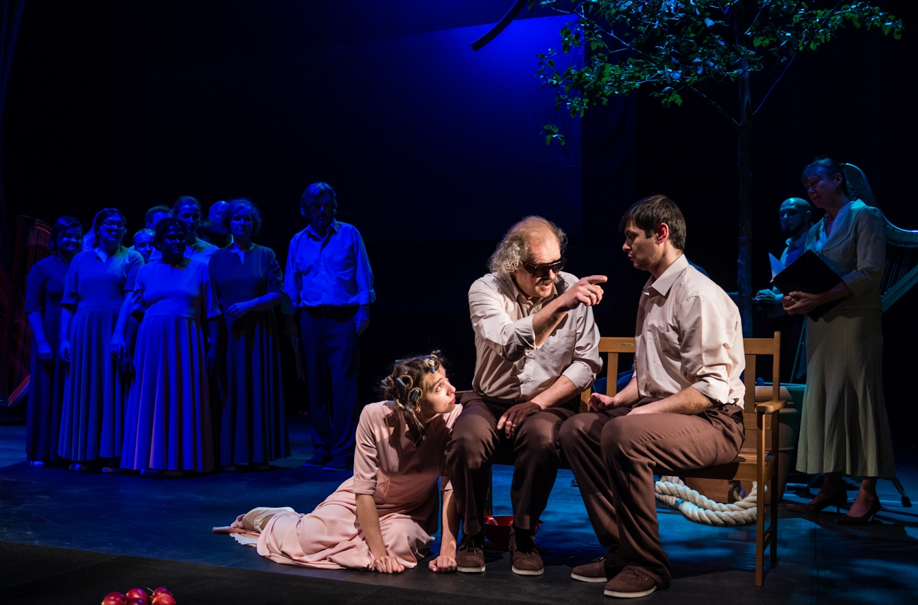 Photograph of a theatre stage. Two male actors are sitting on a bench in the foreground, facing the audience. At their feet is a third actor with curlers in her hair. The actor on the left is wearing dark glasses and pointing towards the actor on the right. In the background, a group of other actors are dimly lit by a dark blue light.