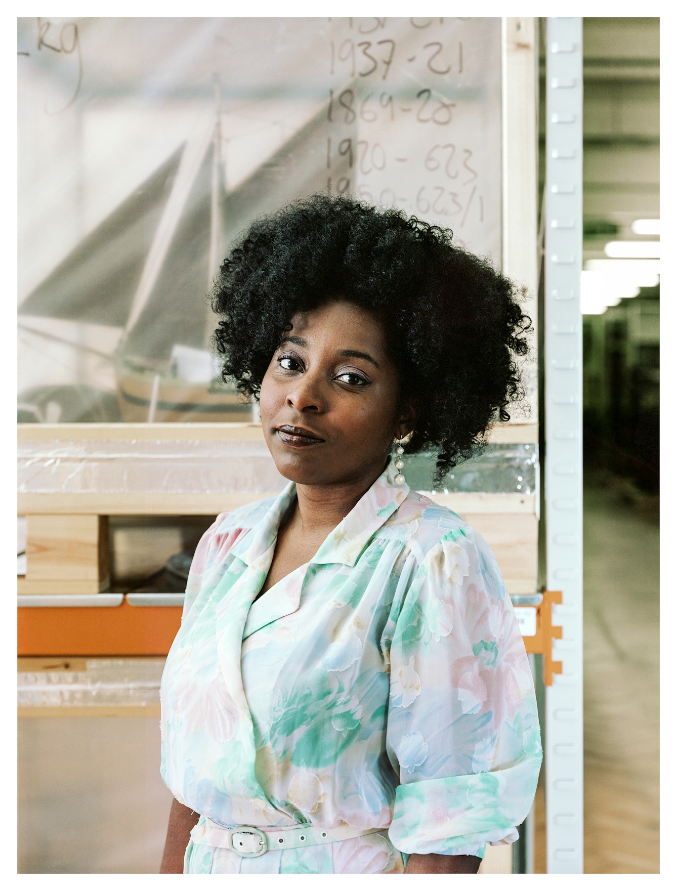 Photographic portrait from the waist up of a woman looking to camera wearing a pastel, floral dress with belt. Behind her is a museum store environment with a large shelving unit. Just visible on the shelf behind translucent plastic sheeting is a ship model, complete with masts and sails.