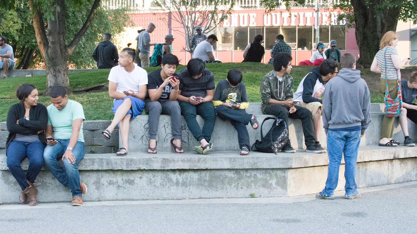 A group of people sitting on a concrete bench in a park, some of them looking intently at mobile phones.