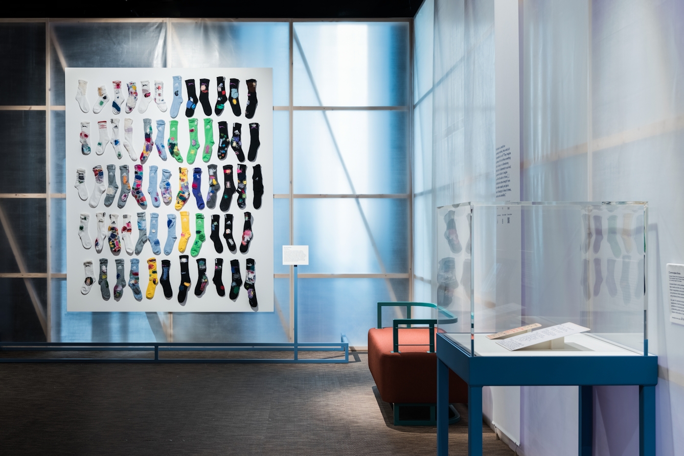 Photograph of an exhibition gallery space showing a translucent fabric backlit wall revealing the wooden structure behind, on which a large rectangular white board is hung. On the front of this board, 60 coloured socks have been mounted in a grid 12 across by 5 high. The tones of these socks transition from light white tones on the left to darker black tones on the right. To the right of the image is another wall at right-angle to the far wall. In front of this wall is a bench seat and a glass display case containing 2 flat paper exhibits.