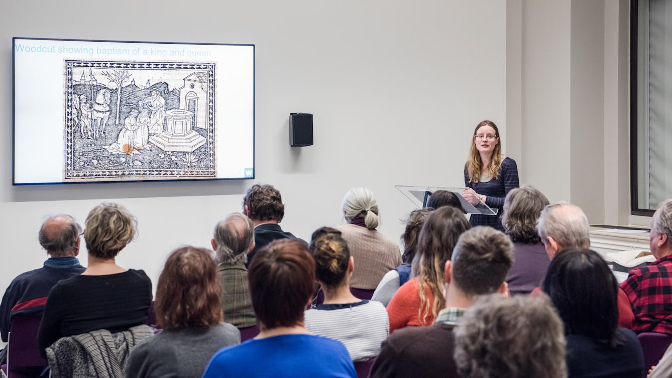 Photograph of a female speaker giving a presentation to an audience. On the wall is a large screen showing an engraving.