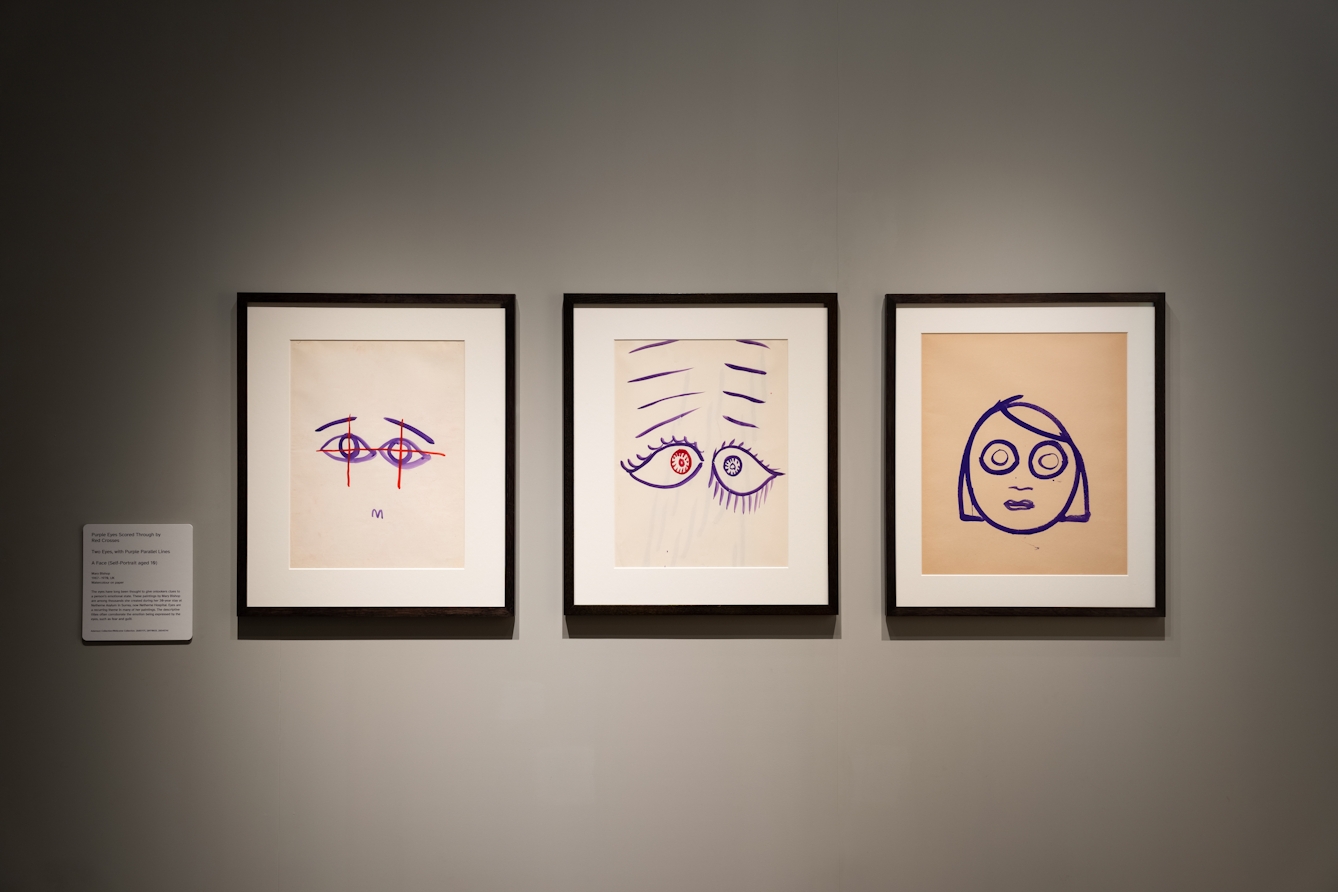 Photograph of 3 framed artworks on a cream coloured gallery wall. Each frame is black edged and contains a hand draw artistic work made up of simple blue and/or red stokes. The work on the left shows a pair of eyes with red crosses through the centre of each pupil. The middle work shows a pair of eyes with eye lashes, where the pupils are crosseyed and there are several parallel lines above each eye. And the work on the right shows la simple face with large circular eyes.