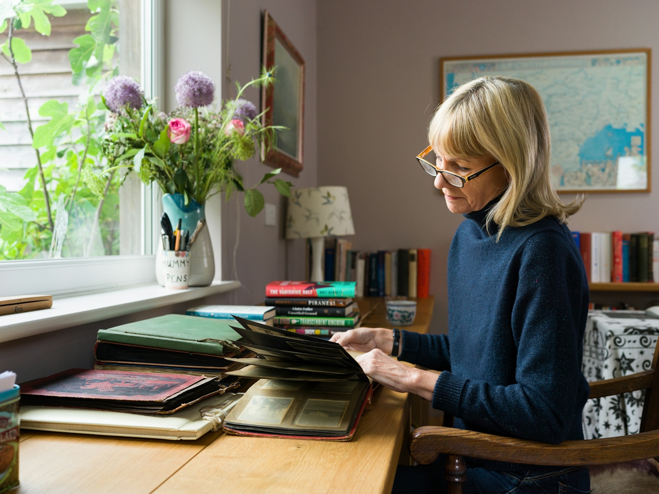 A photograph of a blond women looking through a photo album and sitting on a wooden chair at a desk next to window. On the desk are piles of old photo albums and on the window sill are a flowers arranged in a vase.