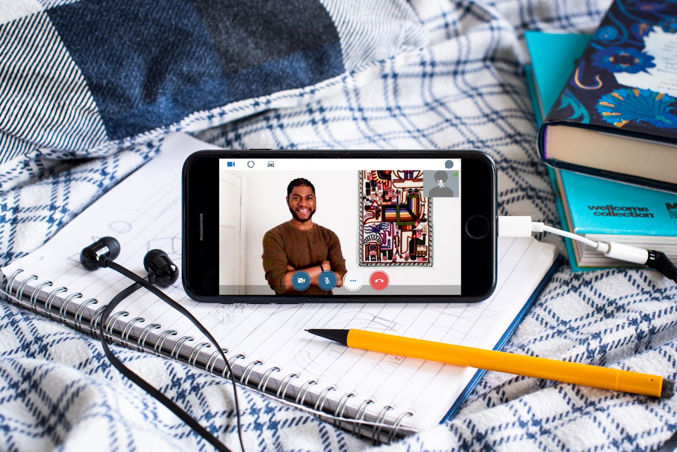 Photograph of a smartphone propped up on a duvet and notebook. Plugged into the phone is a pair of black earbud headphones. On the screen of the phone is a video call with Alain 'Fusion' Clapham who's standing infront of a colourful artwork. Next to the phone are books and a pencil.