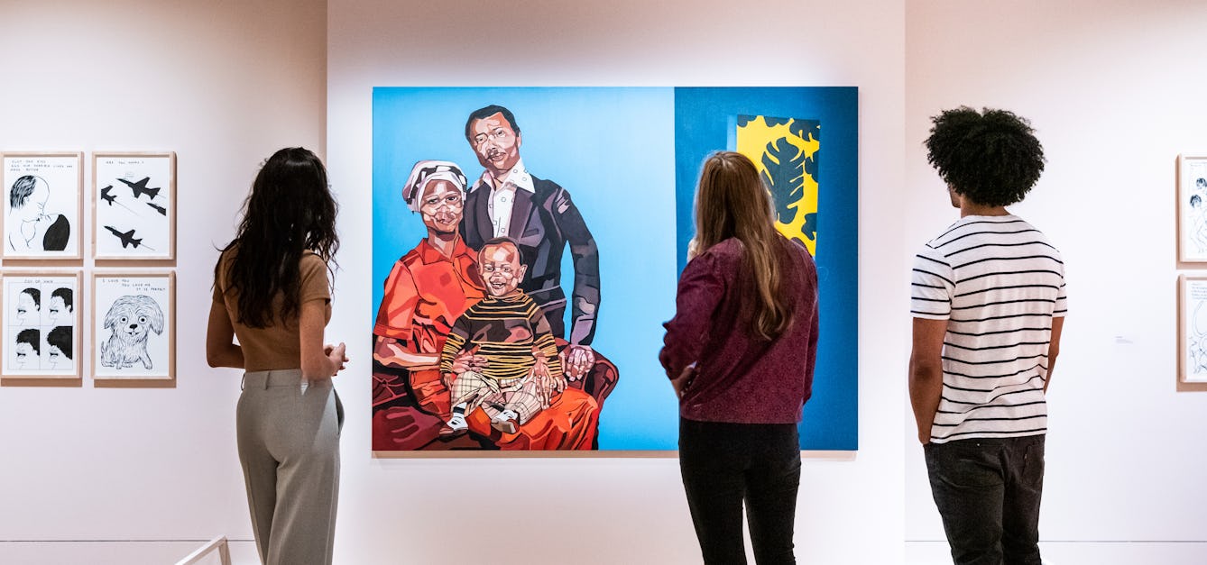 Photograph of an exhibition gallery wall, square on. The wall is a light pink colour and contains a central panel which is set forward from the way, upon which a large colourful old painting is hung. The painting depicts a family portrait with a mother figure a father figure and a young child. At the base of the wall, under the painting is a low white barrier to prevent visitors from getting too close. In front of the painting, standing with their backs to the camera are 3 young people looking at the painting. One stands alone on the left hand side and the other 2 stand together on the right. Either side of the main painting, further small pen and ink artworks can be seen, framed in brown wooden frames.