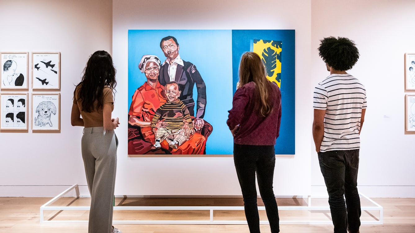 Photograph of an exhibition gallery wall, square on. The wall is a light pink colour and contains a central panel which is set forward from the way, upon which a large colourful old painting is hung. The painting depicts a family portrait with a mother figure a father figure and a young child. At the base of the wall, under the painting is a low white barrier to prevent visitors from getting too close. In front of the painting, standing with their backs to the camera are 3 young people looking at the painting. One stands alone on the left hand side and the other 2 stand together on the right. Either side of the main painting, further small pen and ink artworks can be seen, framed in brown wooden frames.