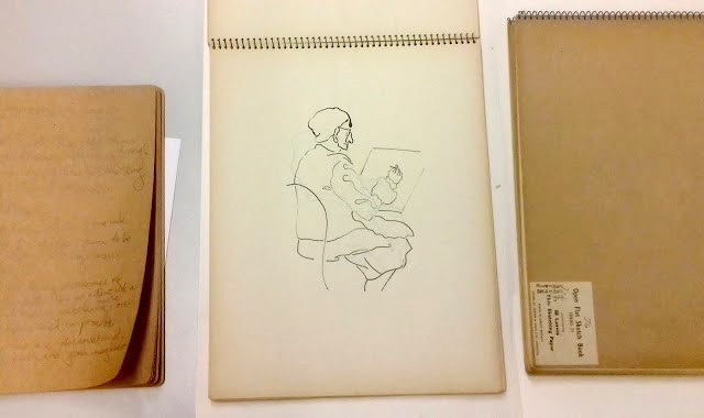 A hand-written notebook and two spiral-bound sketch pads. The central pad is open and has sketch of a seated man drawing.