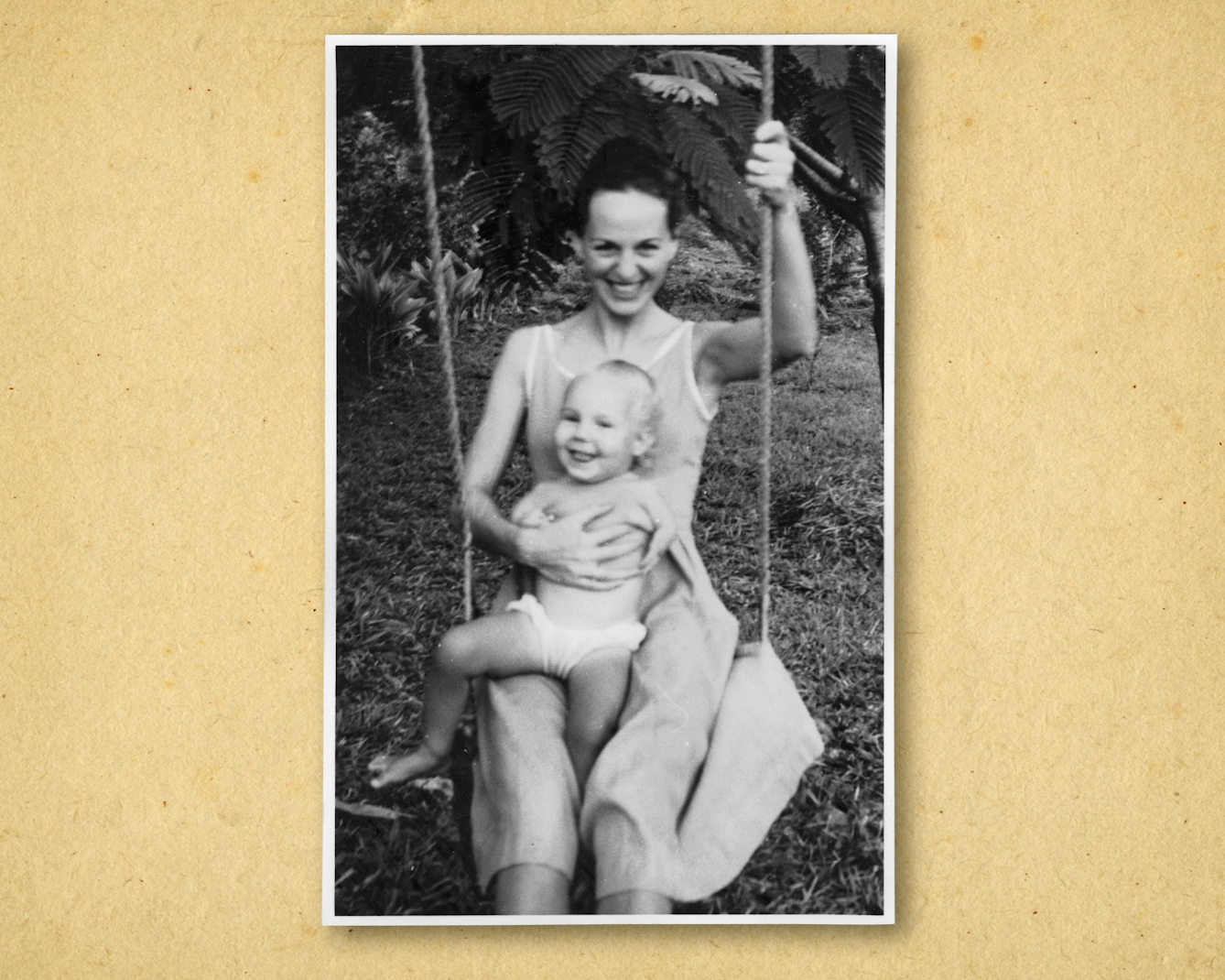 Photograph of a black and white photographic print, resting on a brown paper textured background. The print shows a woman sitting on a swing in a garden. Sitting on her lap is a young child wearing only a nappy. The woman is holding the child with her right arm and holding the swing rope with the other. Both are smiling happily to the camera.