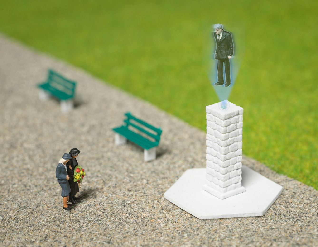 Photograph of model railway scenery depicting a park scene with grass and a wide path. There are two benches and a tall white brick monument. A model of two small figures holding a wreath stand in front of the monument. Out of the top of the monument is a projected holographic image of a man in a suit and tie, looking towards the figures below.