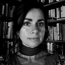 Black and white photograph of the head and shoulders of a young white woman looking straight at the camera. Behind her are bookshelves full of books.