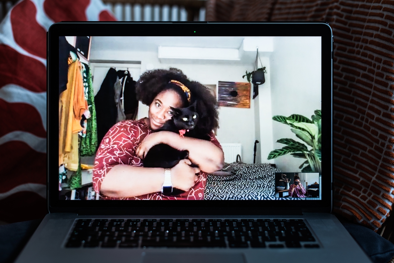 Photograph of an open laptop within a domestic scene. Most of the image is taken up with the screen, with part of the keyboard and trackpad visible. On the screen is a video call showing a woman sitting with a cat in her arms. In the bottom right corner of the screen the photographer can be seen in a small floating window, camera to her eye, in the process of taking the picture.