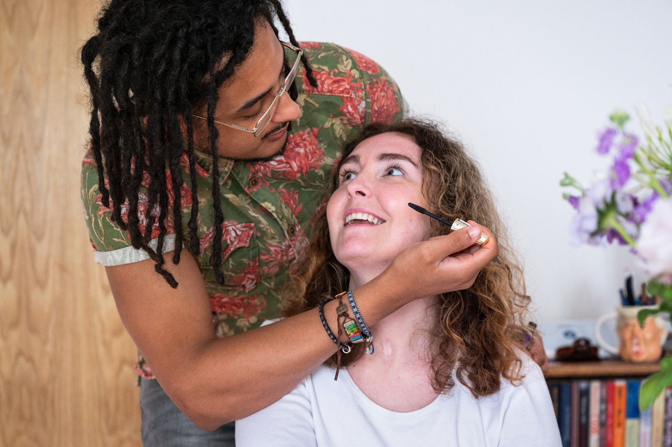 Photograph of a man and a woman inside. The woman is seated and is looking up at the man who is stood behind her, leaning over her. His right arm is wrapped around her, holding a mascara brush near her cheek as if he is about to apply the make-up to her face. He is looking directly at her with a concentrated expression. She is look at him with a loving smile and a hint of nervousness. Behind them both is a plain white wall, a section of a bookcase with objects and flowers on top, and a section of wooden door.