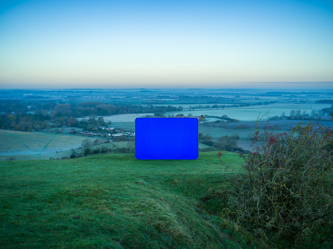 Colour landscape photograph showing a hilltop landscape. The photograph has been taken at dawn and the landscape has a frosty feel to it. In the centre of the image is a large rectangular photographic background frame. Stretched across the frame is a vibrant blue chroma key fabric. The frame is standing on a grass mound on the top of a hill. Behind the screen is the view to the far distance containing fields, tiny buildings and then the horizon. The sky above the horizon is an attractive blend of hues, from warm golden tones at the horizon line blending to a vibrant blue tone as the sky extends upwards. In the foreground to the right is an untidy twiggy shrub, nestled in a hollow.