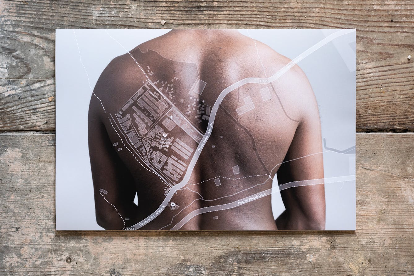 Photograph of a photographic print on a weathered, paint spattered wooden floor. The print shows the back and shoulders of a naked human, against a white background. Superimposed on back and shoulders is a simple line map in white, showing roadways and buildings.
