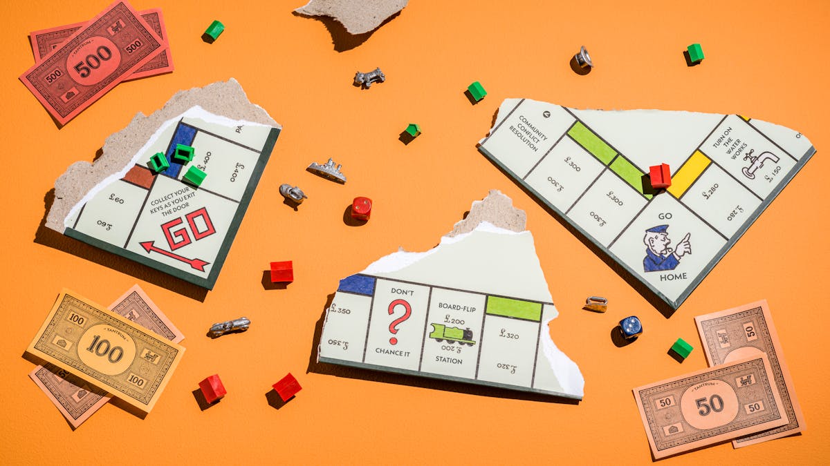 Photograph of a ripped up board game where three board pieces are visible, facing upwards.  scattered around the orange table are pieces of the board game, including  green houses, red hotels, the board pieces and some fake money.  The game title in the money reads "Tantrum".  Other squares on the board game read "Collect your keys as you exit the door", "don't chance it", "board-flip station", "community conflict resolution", and "turn on the water works".