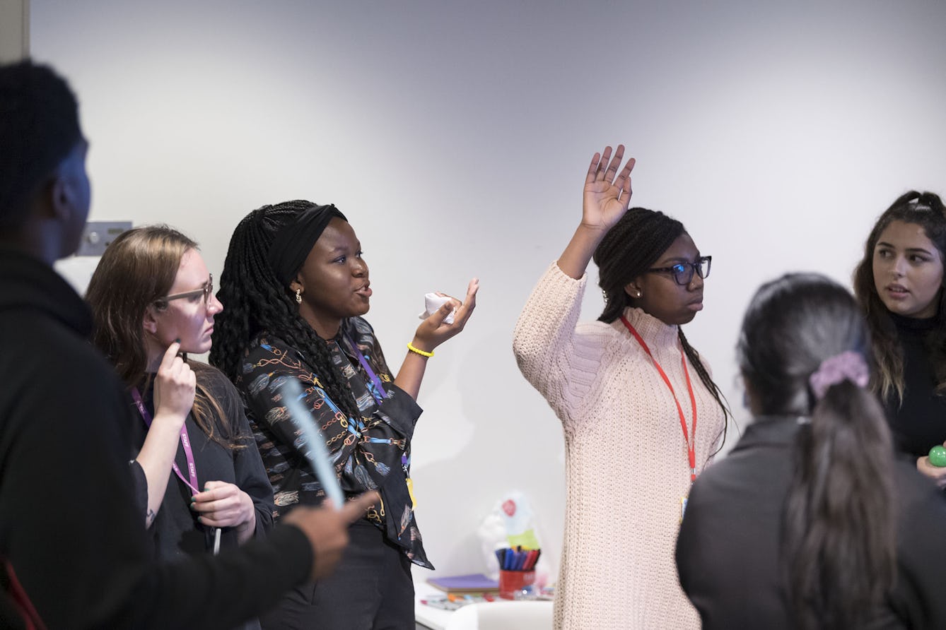 A group of young people having a discussion. One girl has her hand raised, others are looking at another girl, who is speaking.