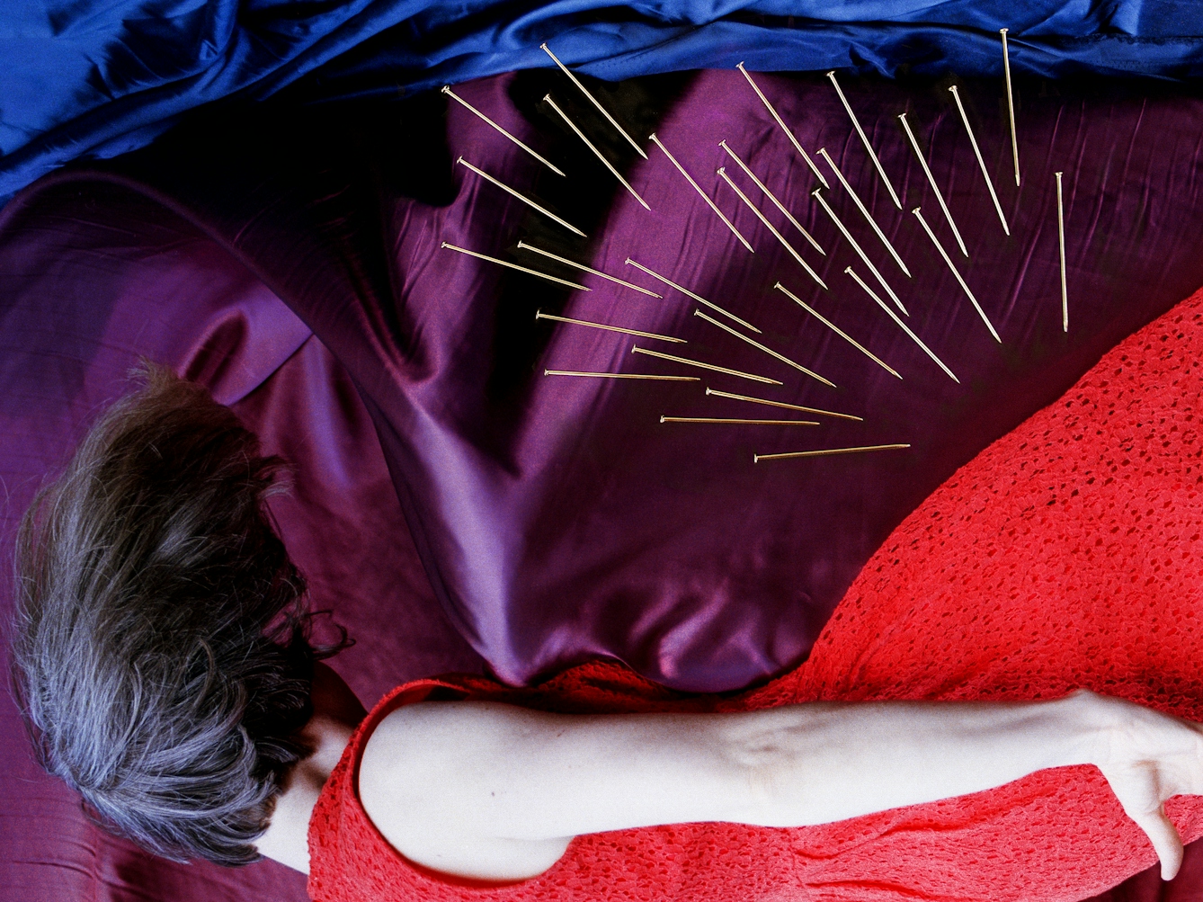 Detail from a larger artwork created with a colour photographic print of a female figure in a bright red dress, set against a purple and blue draped silk background. She is lying horizontal in the frame. Her face is obscured by her hair. Her body is surrounded by groups of dress pins, laid on top of the photographic print. The pins are arranged as if they are a flight of arrows directed at her body. One group attacks her back from underneath.