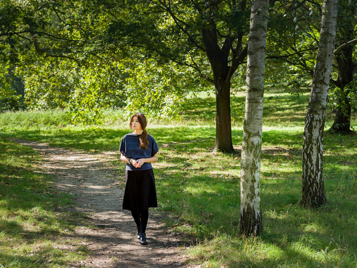 Photographic full length portrait of a woman outside in a woodland scene. She is walking along a rough footpath towards the camera, fingers loosely clasped together. She is looking off to the left. To her left are a series of tree trunks and surrounding the path and the in the background is the green of the grass and leaves of the trees.