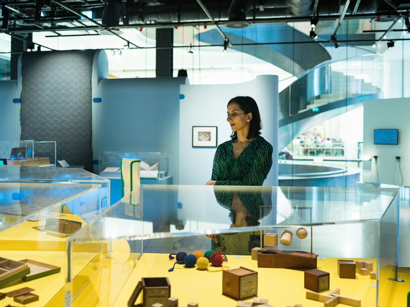 Photograph of a woman in an exhibition gallery space standing behind a glass display case. In the display case are wooden puzzle objects and wooden balls.