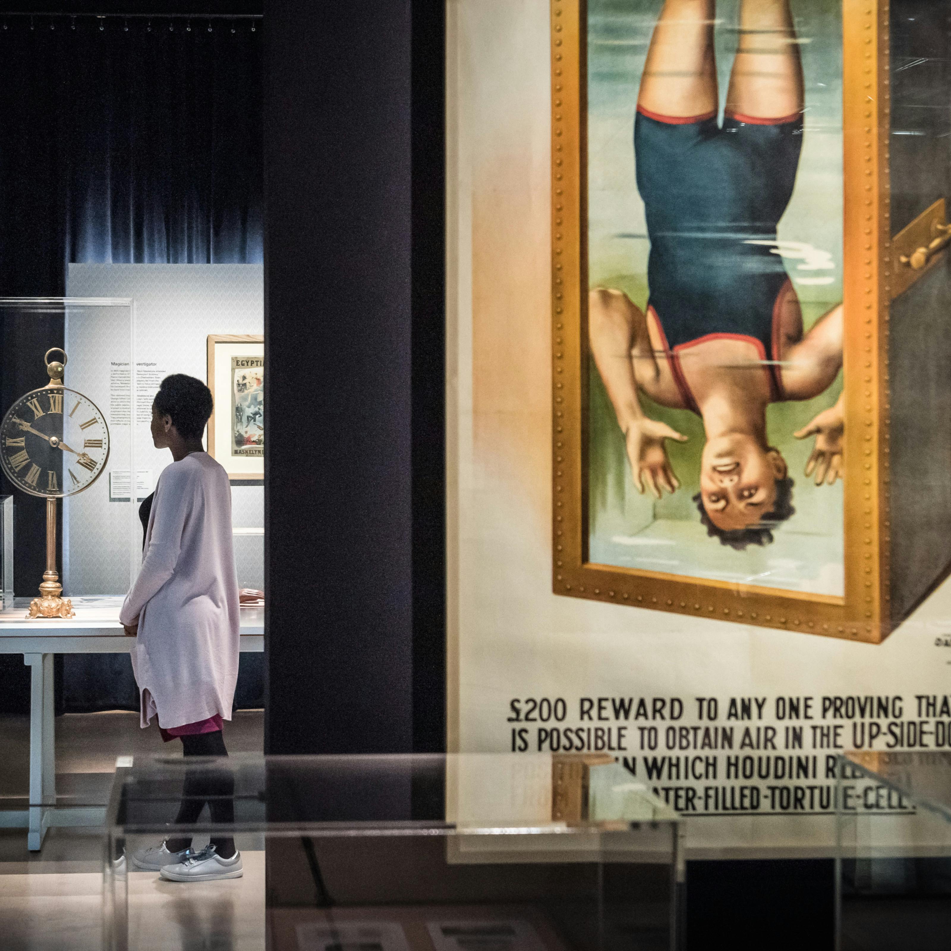Photograph of a visitor exploring the exhibition, Smoke and Mirrors at Wellcome Collection. Photograph shows a women in the background looking at an object in a display case. In the foreground is a large colourful poster showing a man in a Victorian swimming costume suspended upside down in a tank of water.